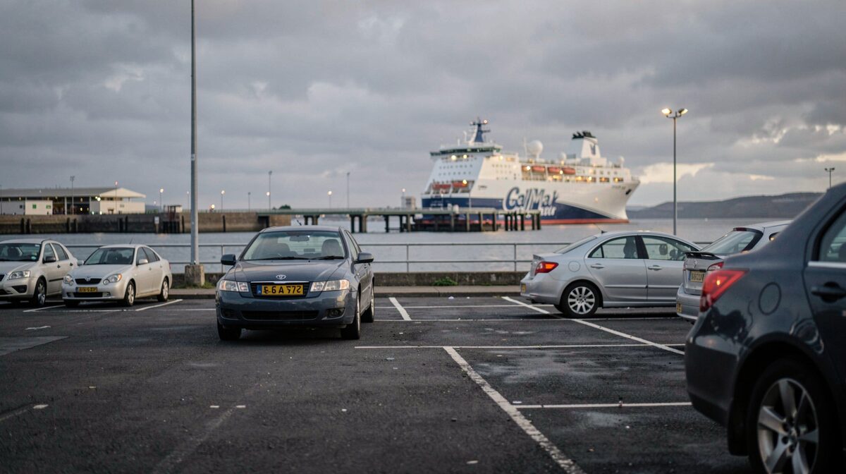 A long-stay car park beside a west coast ferry terminal at first light, one hire car alone under a grey sky, the pier and a CalMac ferry visible beyond it, shot from low behind the car with the number plate out of frame.